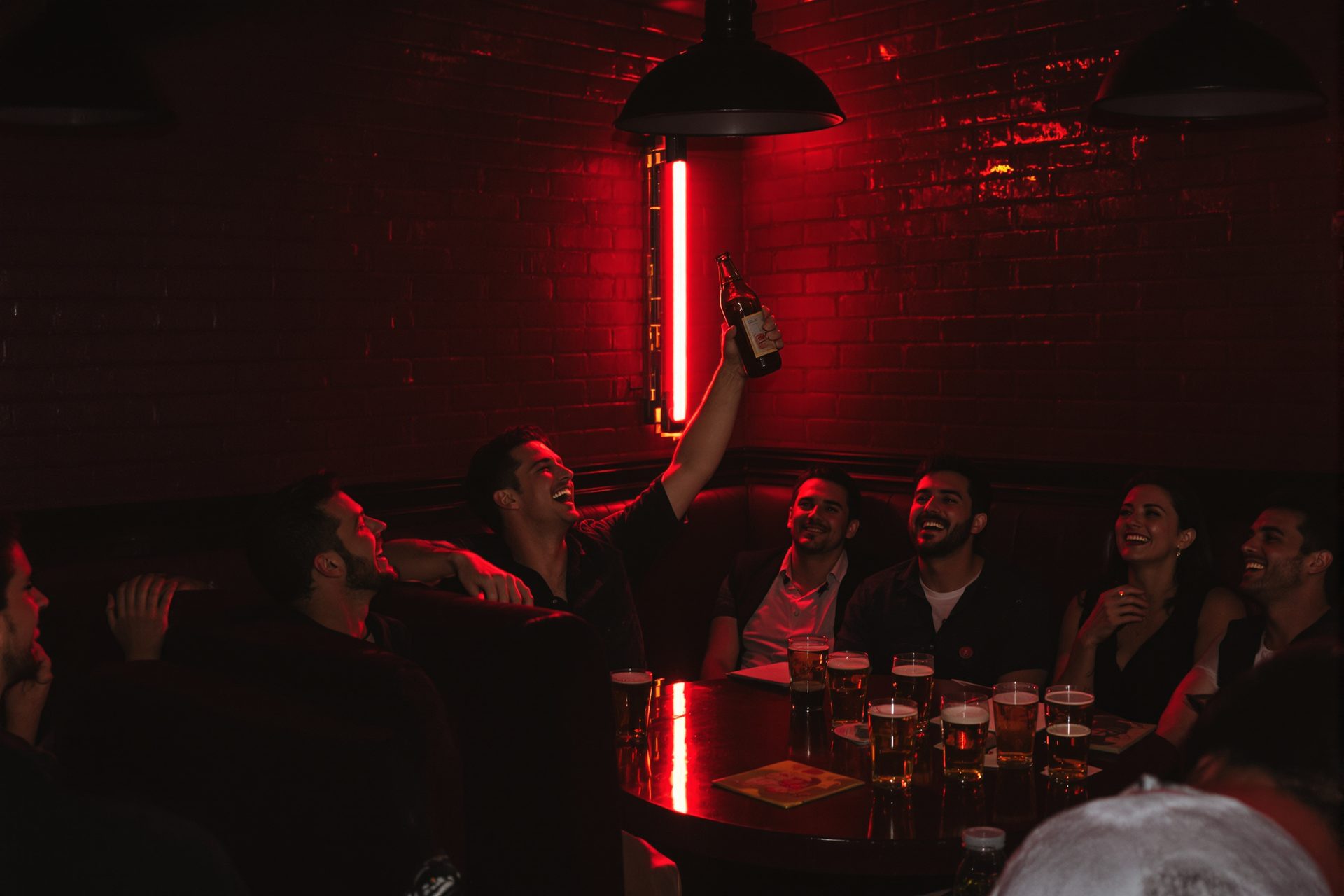 A dim bar at night, one person up out of the booth holding a bottle overhead while the rest of the table laughs.