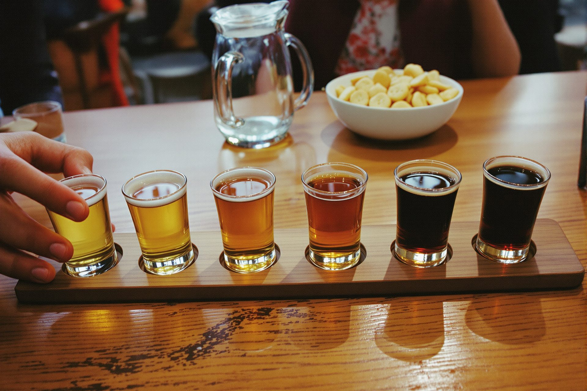 Six small tasting glasses on a bar table, running from pale gold to nearly black, hands reaching in from either side.